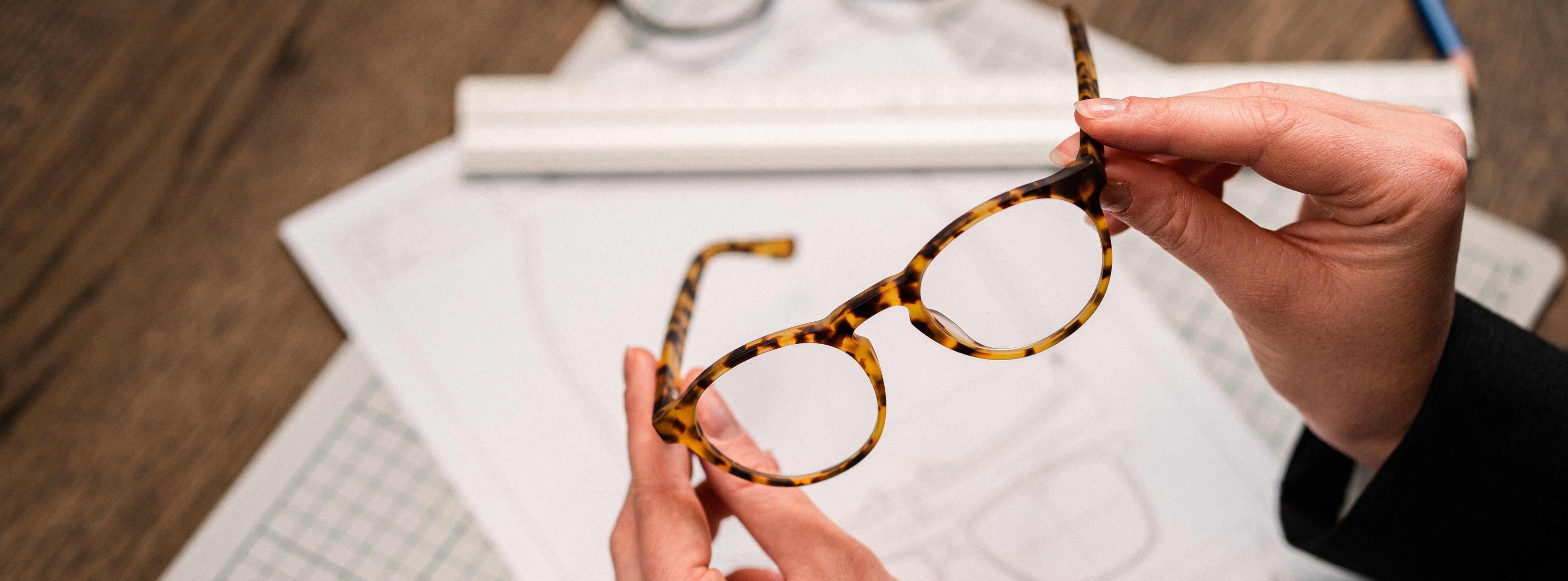 Person holding tortoiseshell glasses over a wooden desk with design papers, highlighting craftsmanship and style.